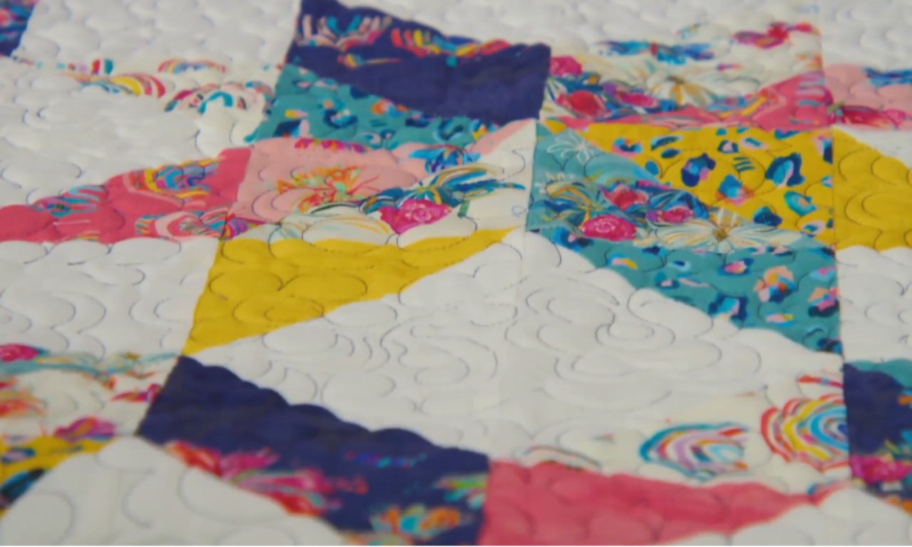 A close-up of a finished quilt top featuring a Carpenter’s Star block. The fabrics are colorful with pink, yellow, blue, teal, and floral patterns. White fabric contrasts with the bright prints, and the quilt is stitched with free-motion quilting in a swirling pattern.