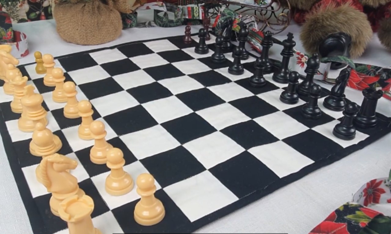 A handmade quilted chessboard with black and white squares laid on a table, set up with cream-colored chess pieces on the left and black chess pieces on the right, ready for a game.