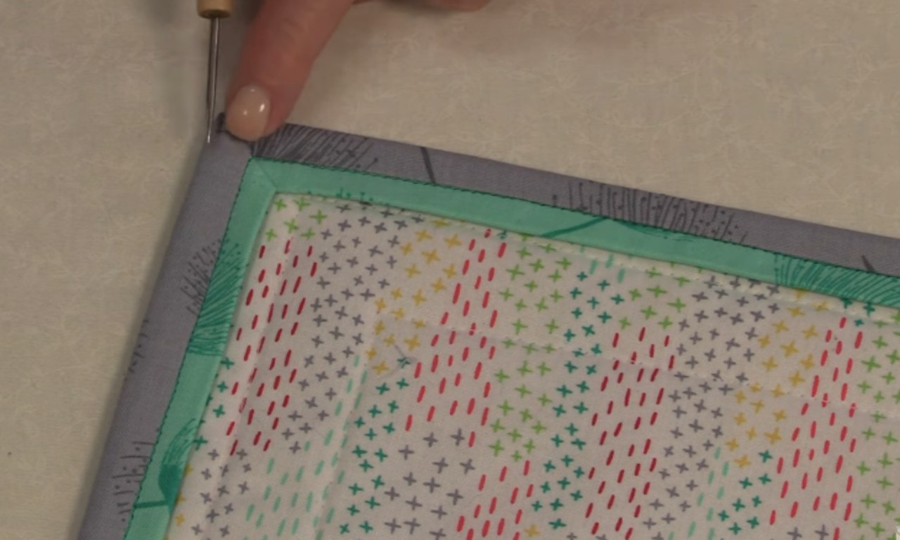 A hand uses a pointed tool to poke out the corner of a quilt, creating a sharp, mitered edge on the gray and turquoise faux flanged binding.