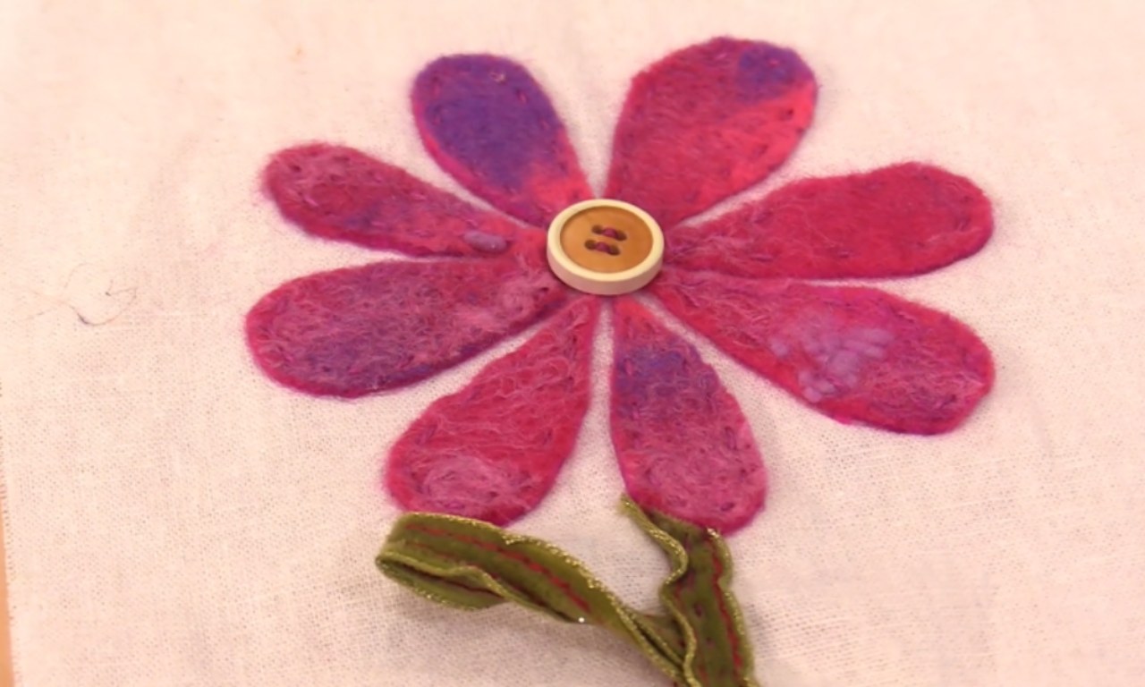 A handmade flower created with felt appliqué on a light fabric background. The flower has mottled pink and purple petals, a wavy green ribbon stem, and a wooden button for its center.