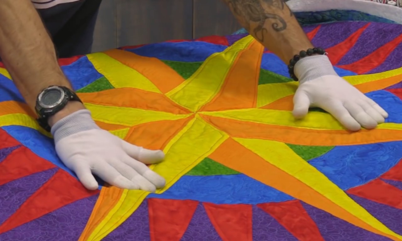 A quilter wearing white gloves smooths down a colorful Mariner's Compass quilt top made of yellow, orange, and blue fabric.