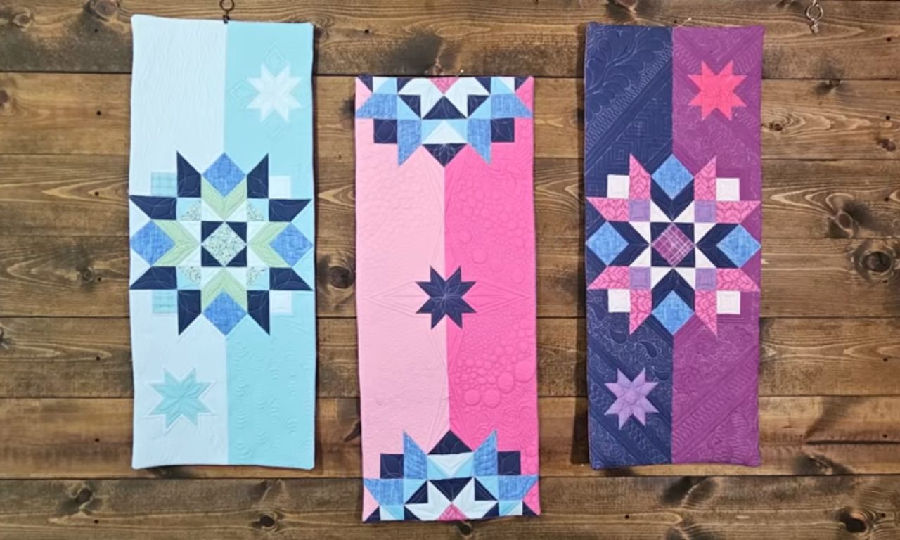 Three quilted wall hangings with colorful star patterns displayed against a wooden background, featuring blue, pink, and purple designs.