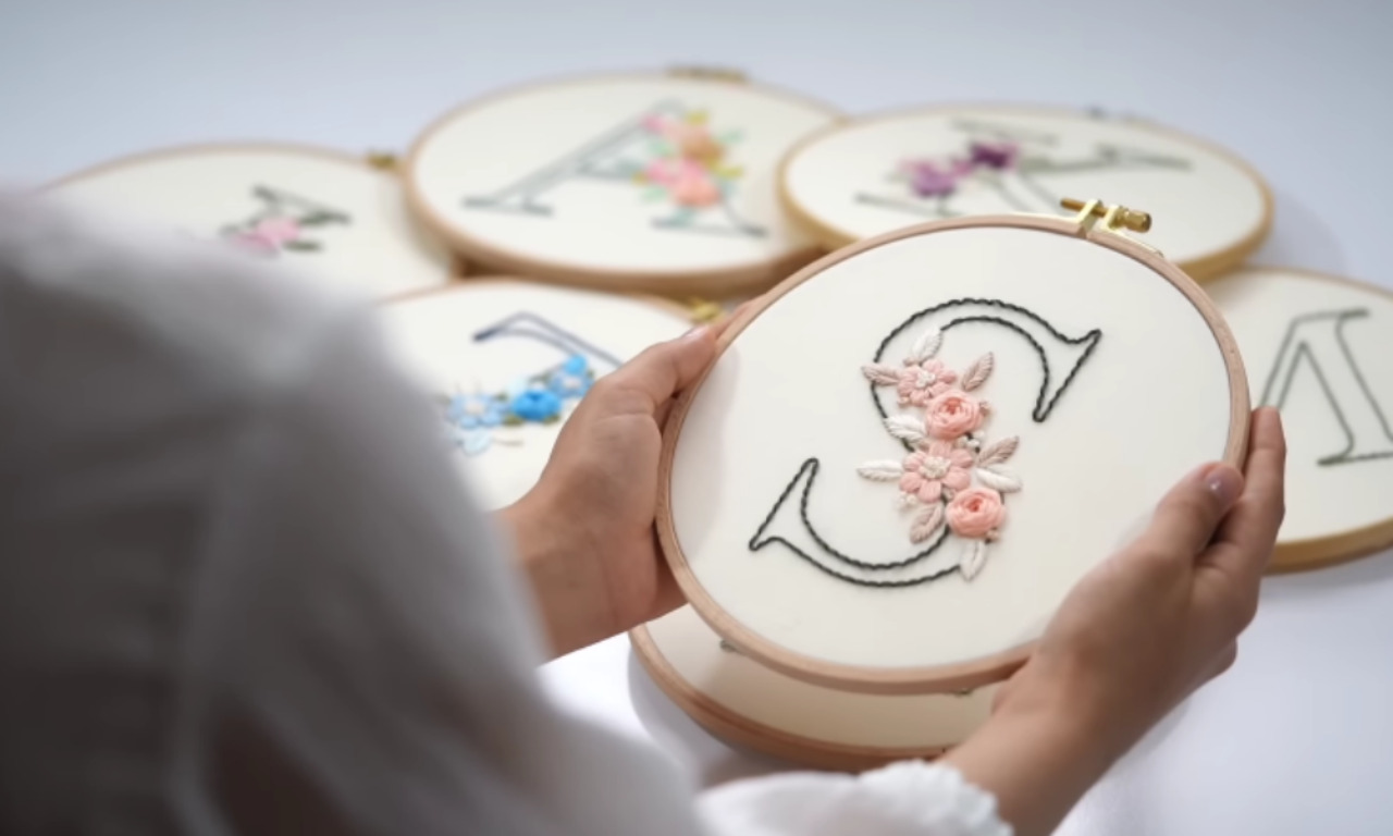 A person holding a finished hand embroidery hoop with the letter S decorated with delicate pink and white flowers, showing a beautiful example of a hand-embroidered monogram.