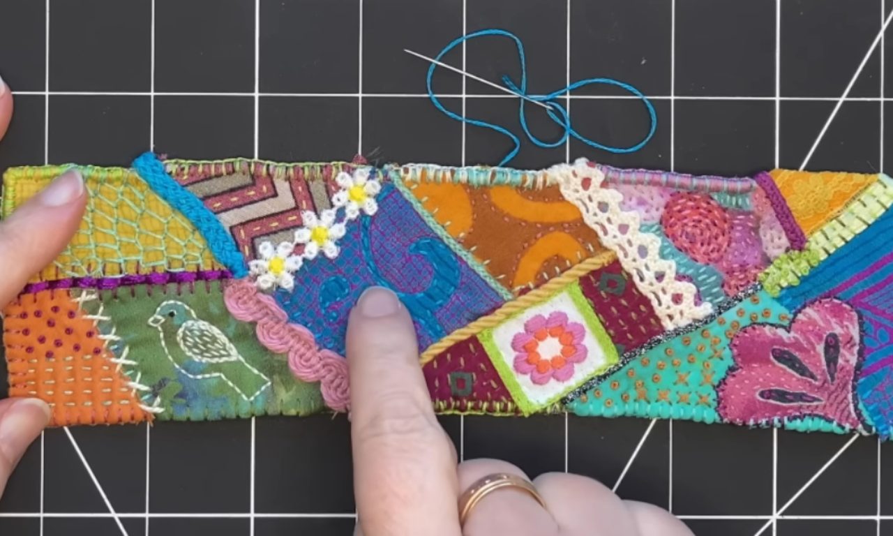 A person's hand pointing to a completed modern crazy quilt strip, showing the rich texture from various fabrics, decorative stitches, and embellished seams with lace and daisies.