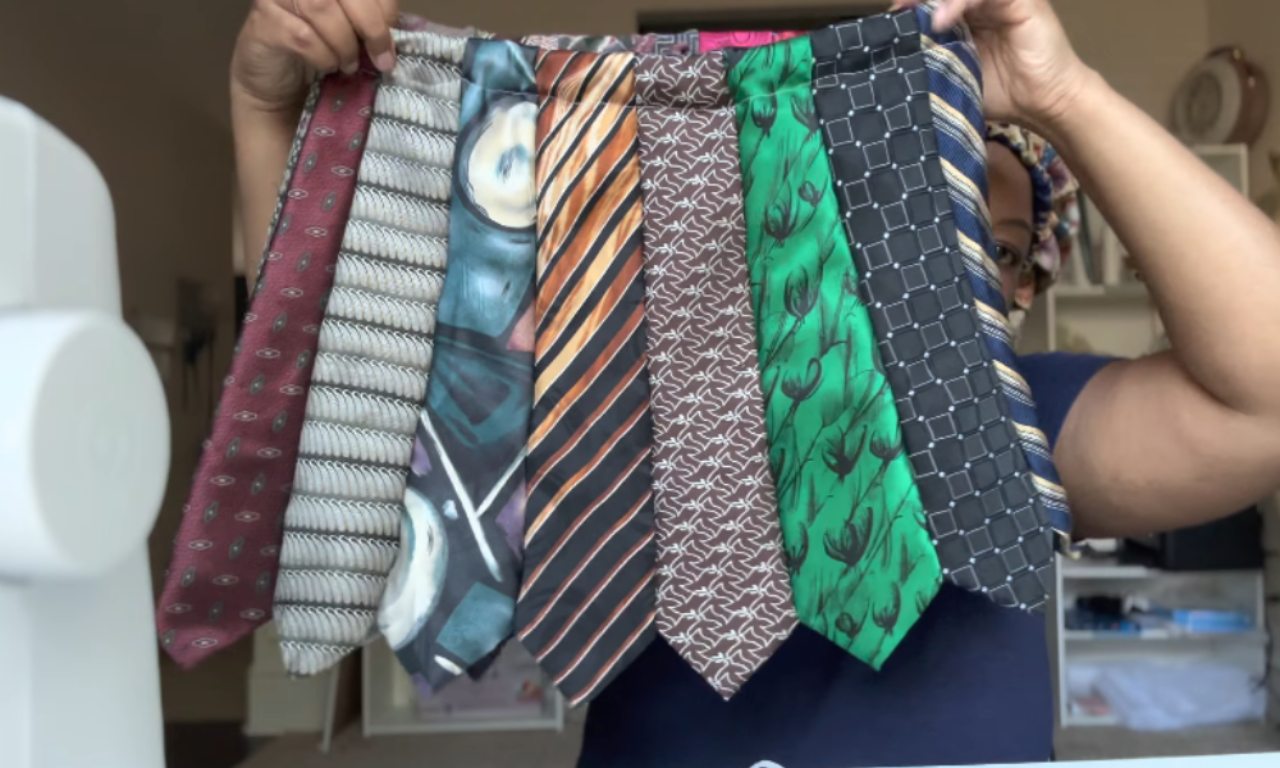 A person holds up several colorful neckties stitched together side by side, forming the beginning of a skirt. The ties display different patterns, including stripes, geometric shapes, and abstract designs.