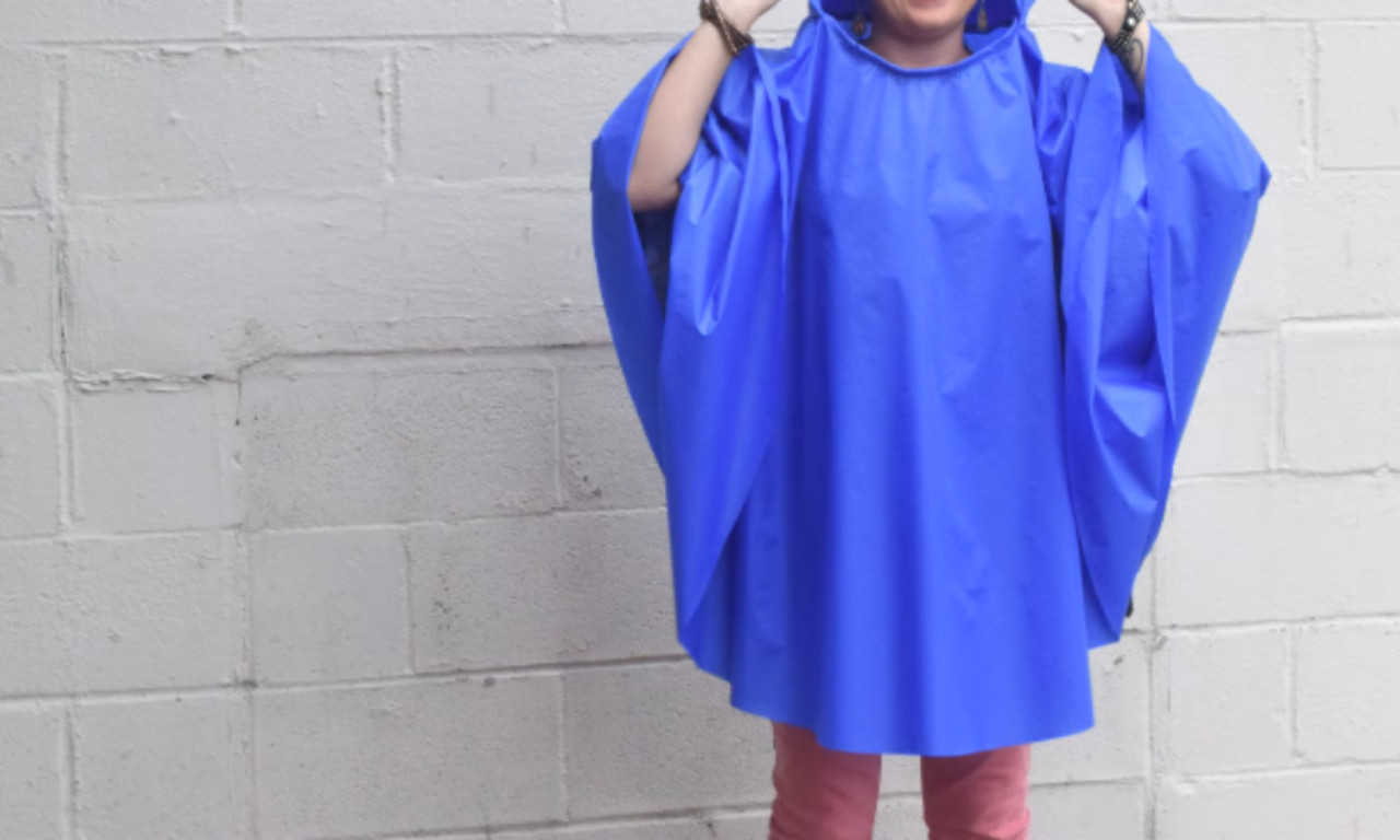 A person holding up a completed bright blue rain poncho, smiling with arms outstretched. The poncho has wide, loose sleeves and a simple design. A plain white brick wall serves as the background.