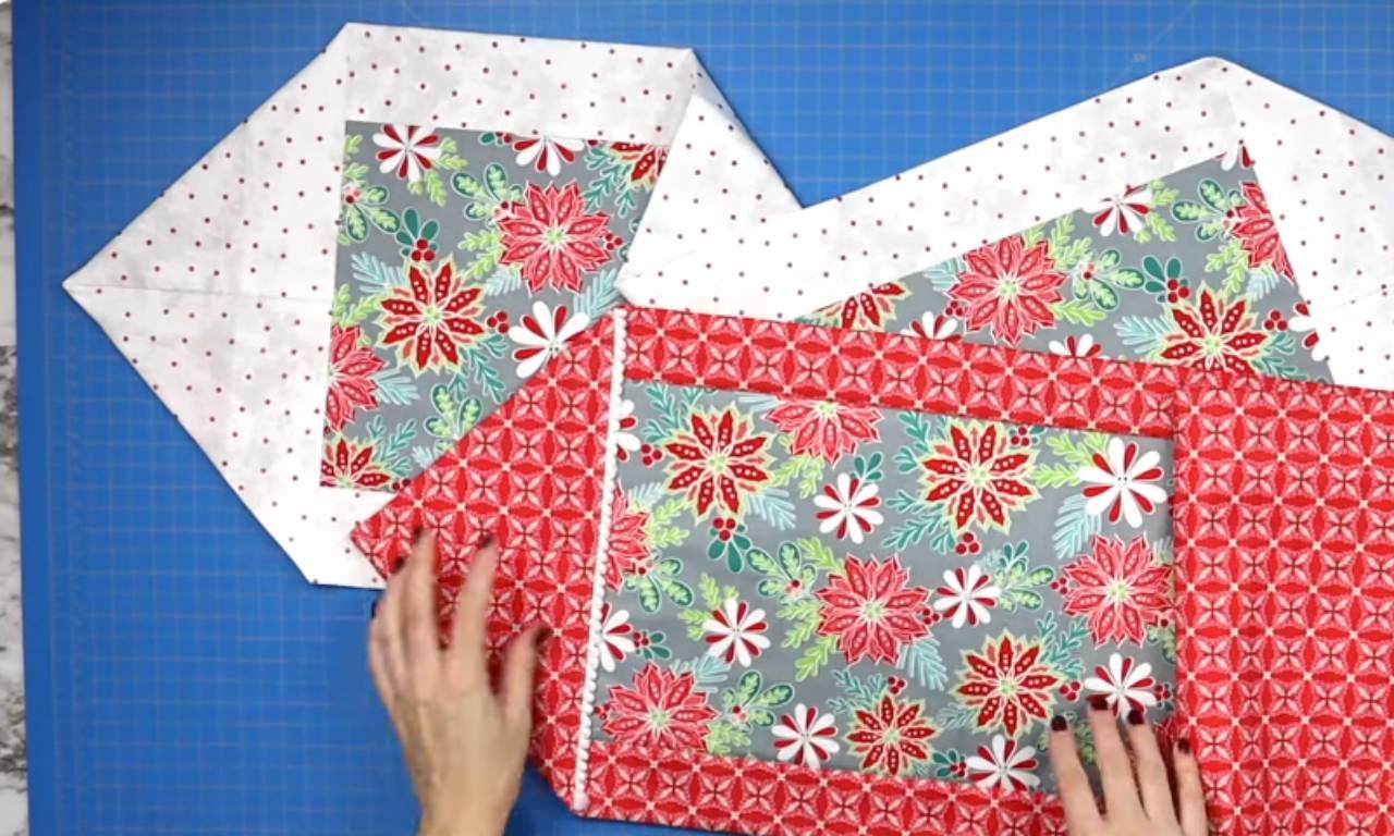 Hands arranging fabric pieces to form a reversible table runner, featuring a festive floral print bordered with red geometric patterns and white fabric with small red dots on the back.