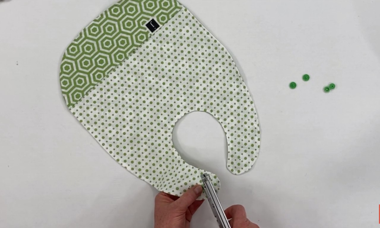 A finished baby bib made from green patterned fabric, with one side polka-dotted and the other hexagon patterned. A person is using a snap fastener tool to attach green snaps to the neckline closure.