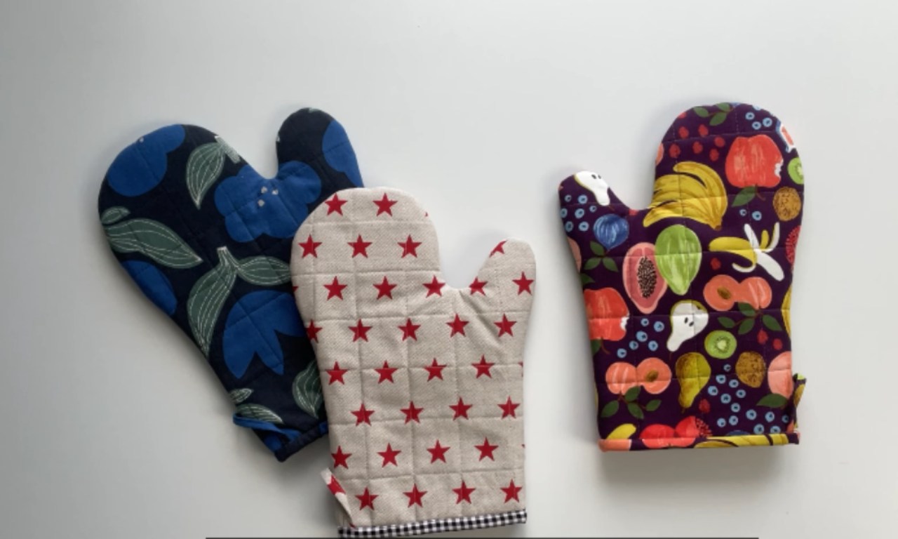 Three finished homemade quilted oven mitts displayed on a white background, each with a unique fabric pattern—floral, stars, and fruit.
