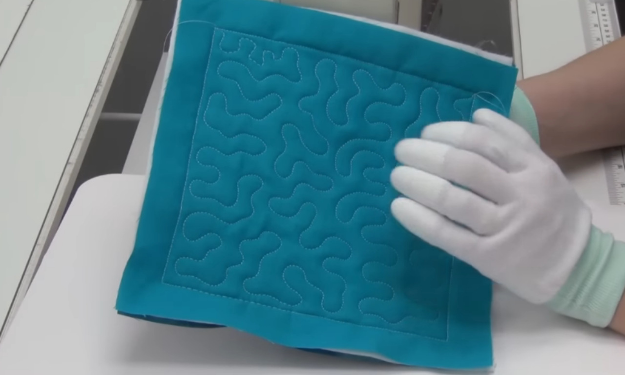 Person wearing white gloves holding up a teal quilt square with free-motion quilting in a meandering pattern.