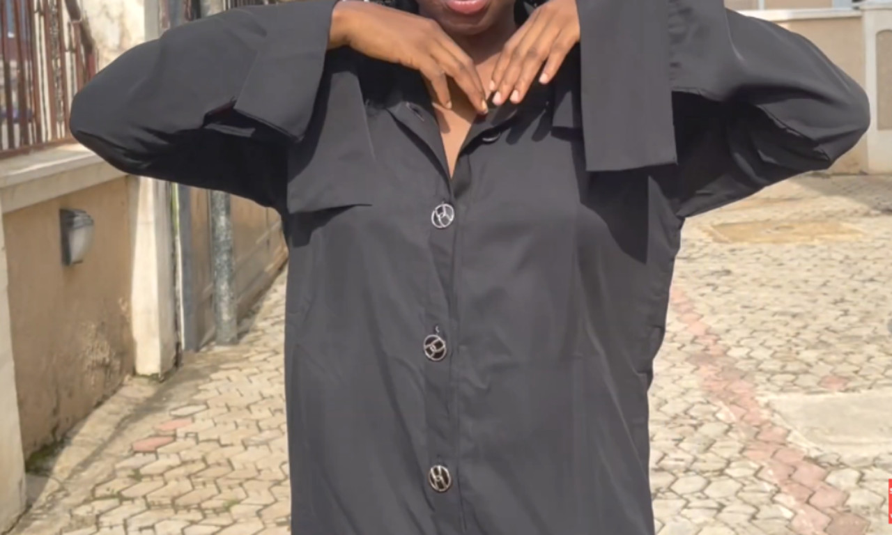 A person wearing a loose-fitting black collared shift dress with oversized sleeves and large round decorative buttons, standing outdoors with arms slightly lifted.