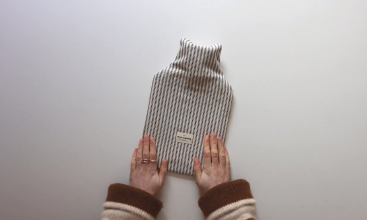 Two hands holding a finished hot water bottle cover made from striped fabric with a small fabric label at the bottom center, placed on a plain white surface.