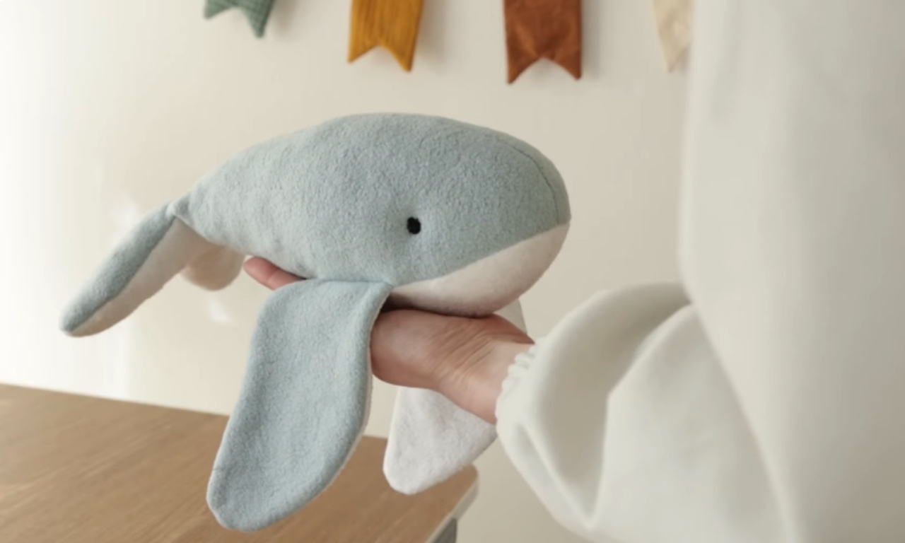 A person holding a handmade plush toy whale made of soft, light blue and white fabric. The whale is held in one hand, and a decorative banner hangs on the wall in the background.