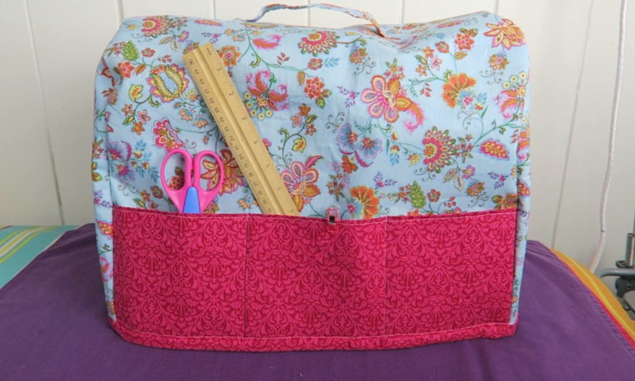 A finished, custom sewing machine cover made of light blue floral fabric with a large, bright pink pocket on the front. The pocket holds a wooden ruler and a pair of pink scissors.