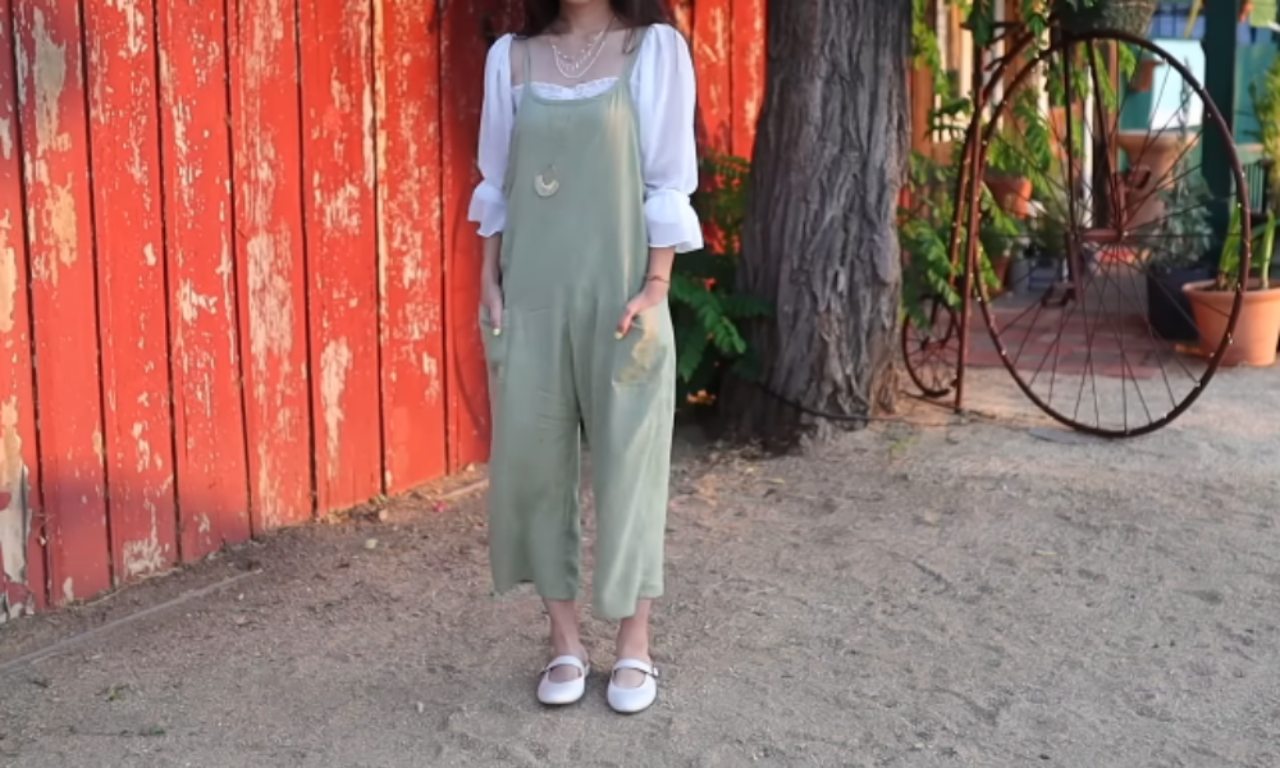 A person standing outdoors wearing a green sleeveless jumpsuit over a white blouse. The outfit is paired with white shoes and accessorized with layered necklaces. The background shows a rustic red wooden wall with some greenery.