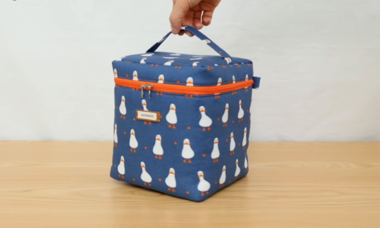 A blue insulated lunch bag with a duck pattern, orange zipper, and a handle on top is shown standing on a wooden surface. A hand is holding the top handle.