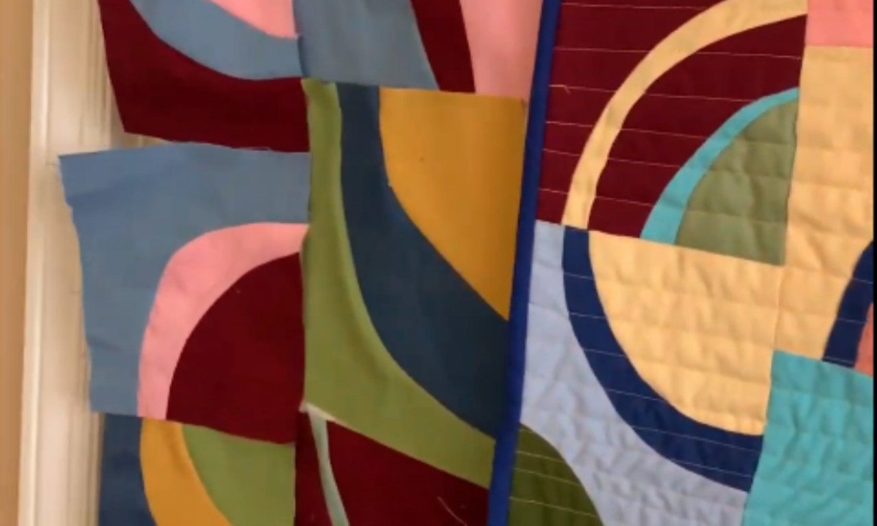 Partially sewn curved quilt blocks pinned beside a finished quilt. The quilt shows bold overlapping curves in burgundy, yellow, green, blue, and pink fabrics with straight-line quilting.