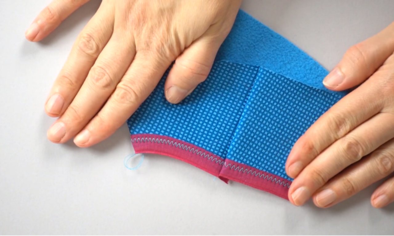 A pair of hands holds a piece of blue fabric, showing a cleanly finished edge with pink fold-over elastic attached by a zigzag stitch.