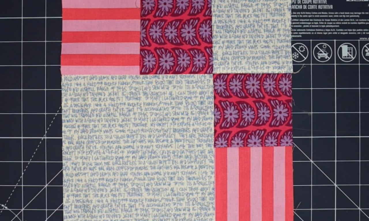 A finished quilt block made of various patterned fabric strips in red, pink, blue, and white colors is laid flat on a black cutting mat with white grid lines.
