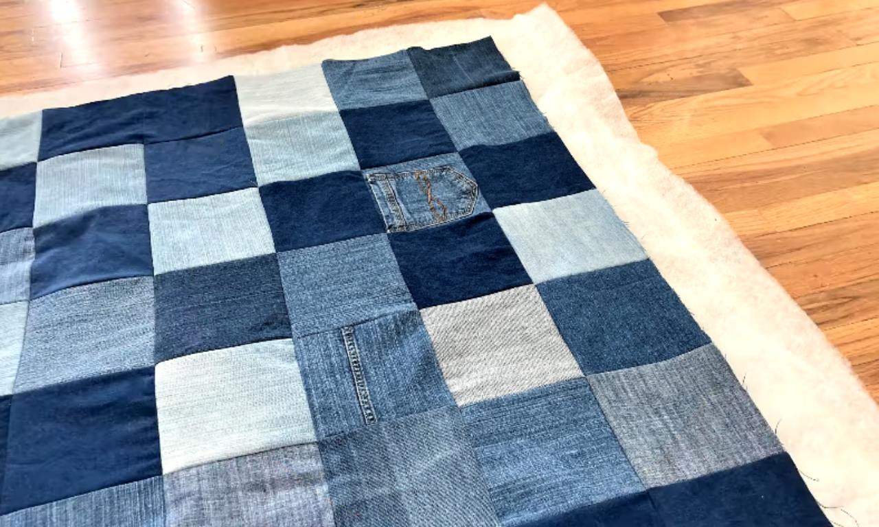 A DIY denim quilt top made of blue jean squares is laid out on white batting on a wood floor, showing the first step of making a quilt sandwich.