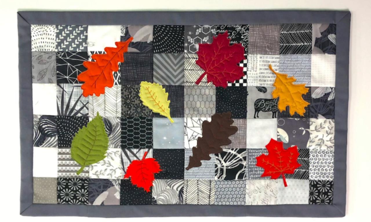 A quilt featuring a variety of black, white, and gray patterned squares with bright-colored felt leaves in red, orange, yellow, and green scattered across the quilt. The quilt has a gray border and appears to represent a seasonal or autumn theme.