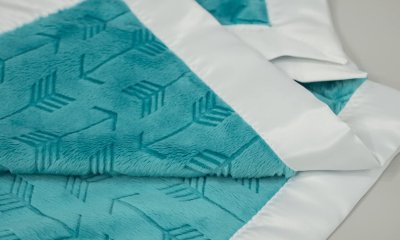 A finished baby blanket made from blue minky fabric with an arrow pattern, bordered with a smooth white satin binding. The blanket is neatly folded to display the corners and edges.