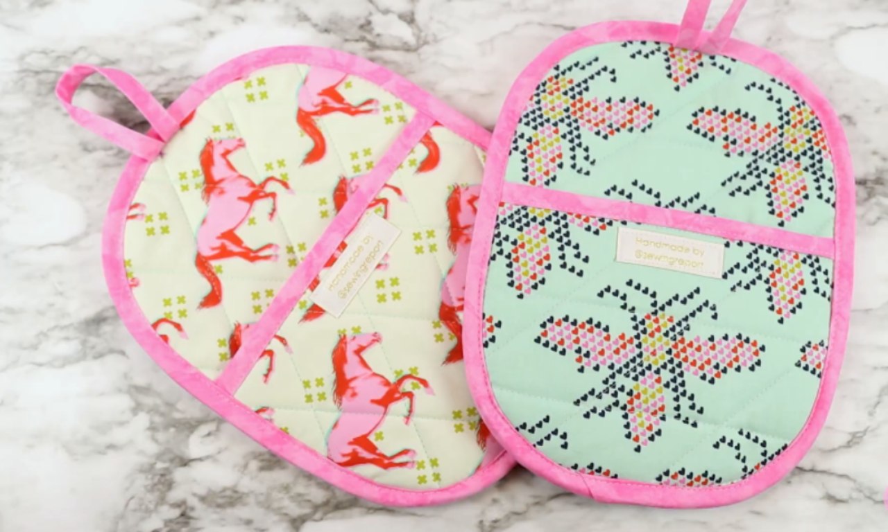 Two finished, oval-shaped pot holders with hand pockets and pink binding, displayed on a marble background.