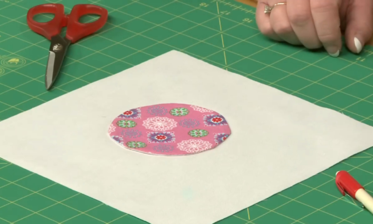 An oval piece of pink floral fabric is centered on a square of white fusible interfacing, sitting on a green cutting mat next to a pair of red scissors.