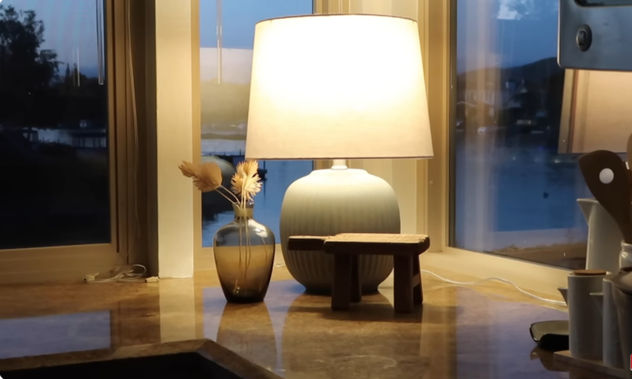 A close-up shot of a kitchen counter at dusk, featuring a modern table lamp with a soft white shade and a pale blue ceramic base. Next to it is a small glass vase with a dried floral arrangement. The lamp's warm light is reflected on the glossy countertop and the window behind it, which overlooks a body of water and hills.