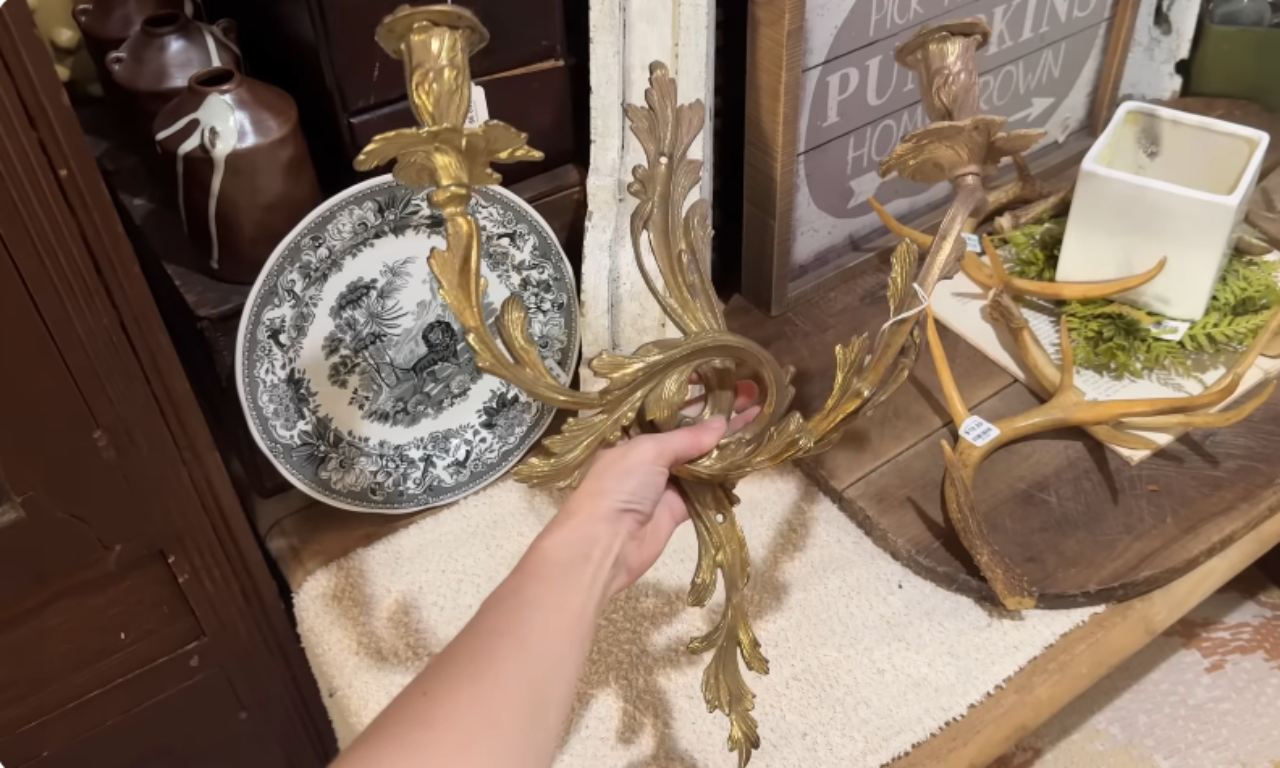 A close-up view of a hand holding a large, ornate, antique brass wall sconce with leaf and scroll details, set against a background of a decorative plate, antlers, and rustic wooden surfaces.