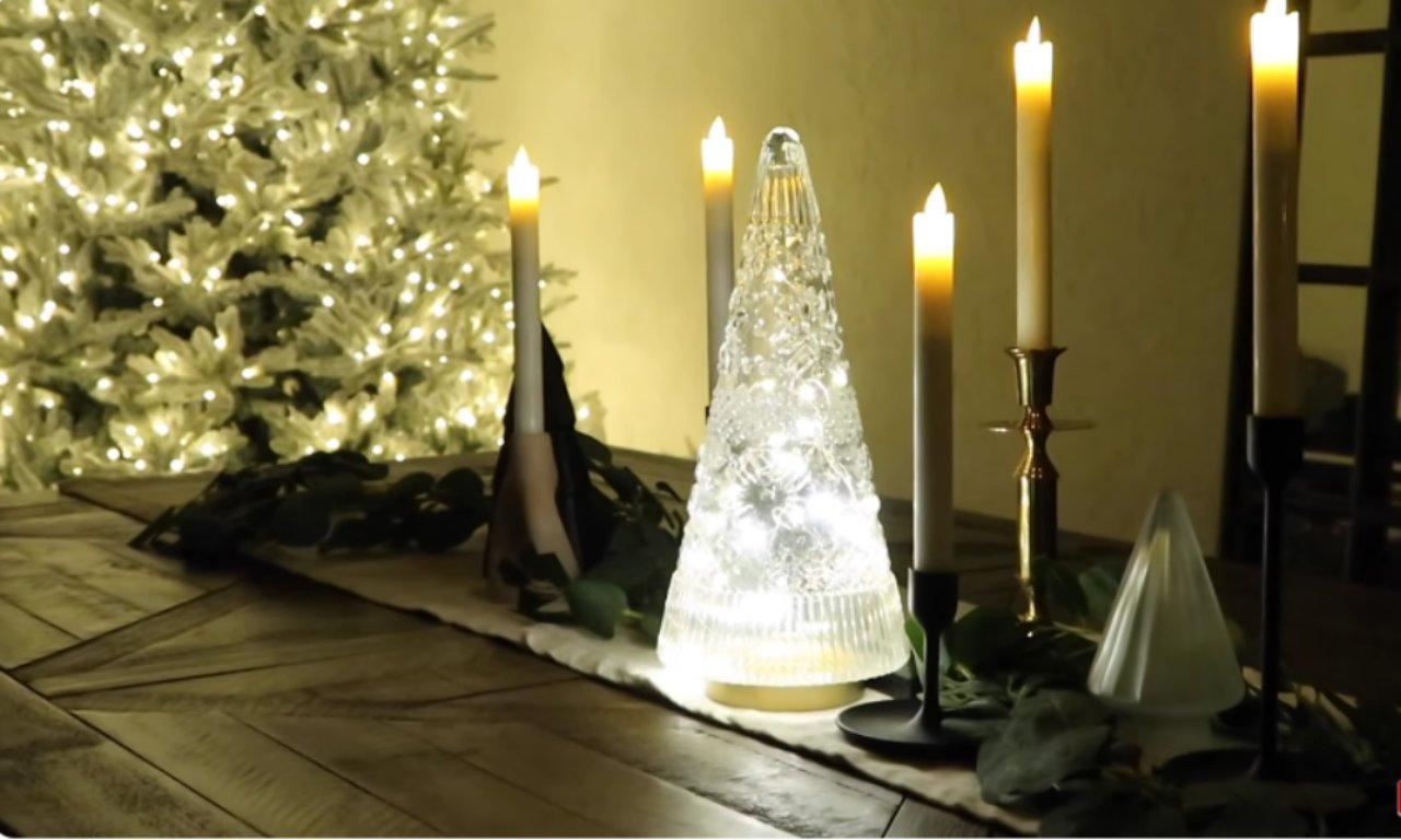 A wooden table is set with a white and black runner. An assortment of flameless candles and decorative light-up trees sits on top. A flocked and lit Christmas tree is visible in the background.