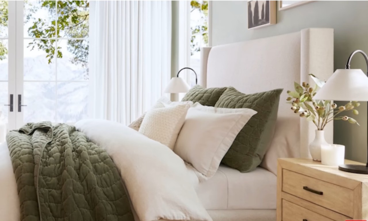 A close-up view of a cozy, well-lit bedroom. A large, plush bed with a cream-colored wingback headboard is covered in crisp white and quilted olive green bedding. White accent pillows rest on the bed. A rustic wood nightstand holds a white vase with eucalyptus sprigs and two white candles. A modern black table lamp illuminates the scene. Large windows with sheer white curtains are visible in the background.