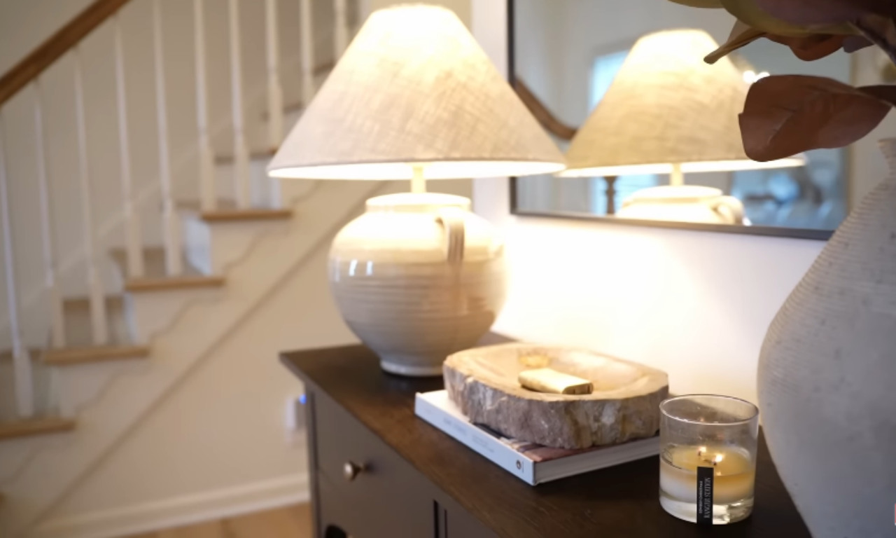 Close-up of a dark wood console table in an entryway, featuring a ceramic jug lamp with a linen shade, a lit scented candle in a glass jar, and a small wooden bowl with a natural, rustic edge sitting on top of a book. A white staircase with natural wood treads is visible in the background to the left, and a mirror reflects the lamp on the wall to the right.