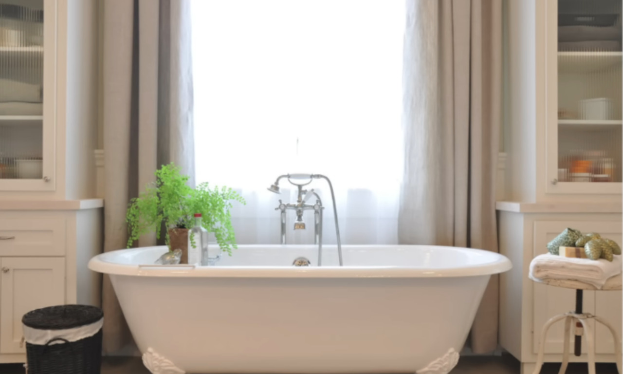 Bright bathroom with a freestanding white clawfoot bathtub, centered under a large window with sheer curtains, surrounded by white cabinetry and decorative plants.