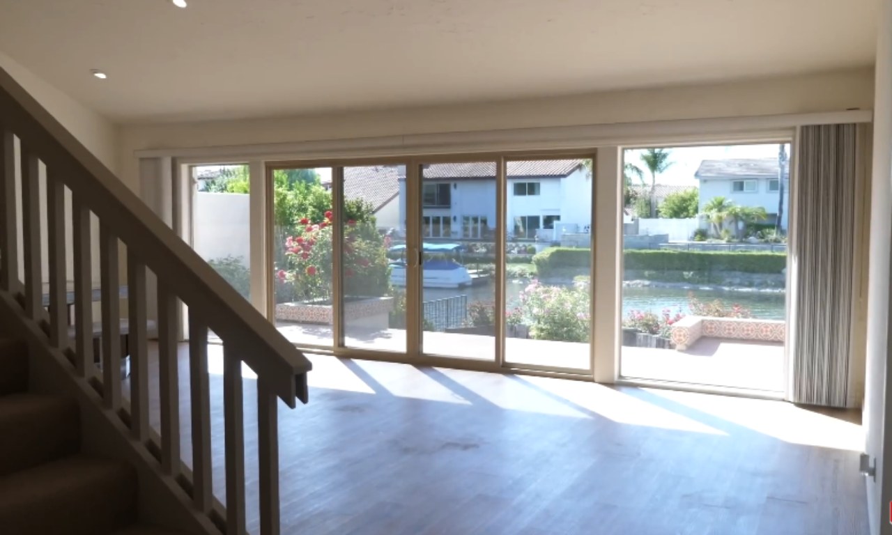 An empty interior room with wooden flooring and a staircase leading up on the left. The room is dominated by large glass sliding doors and windows that open onto a sunny backyard with a waterway, showing lush green foliage and houses across the canal.