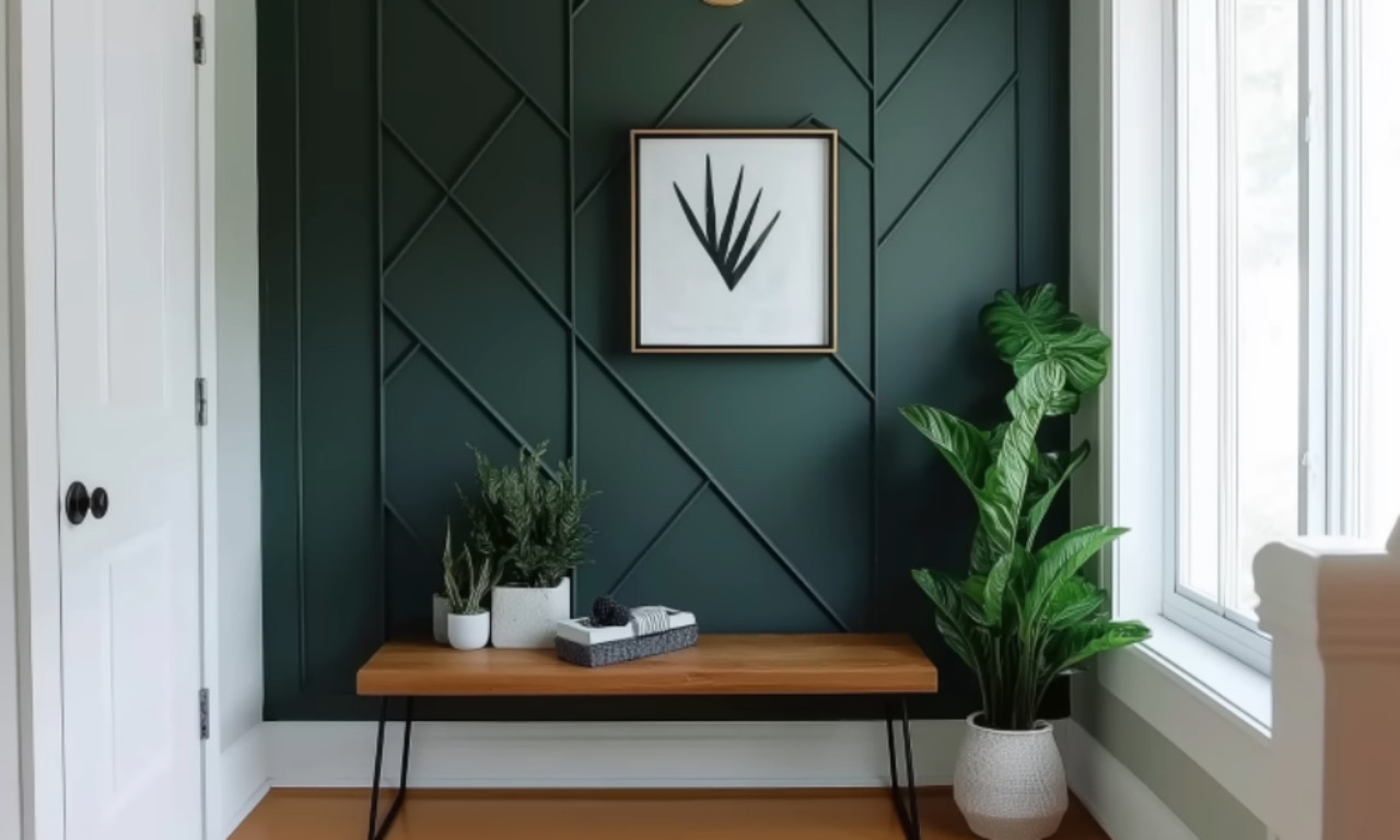A modern entryway featuring a dark green accent wall with geometric paneling, a wooden bench with black metal legs, and potted plants. A minimalist black and white framed artwork is centered on the wall above the bench. Natural light enters from a large window on the right.