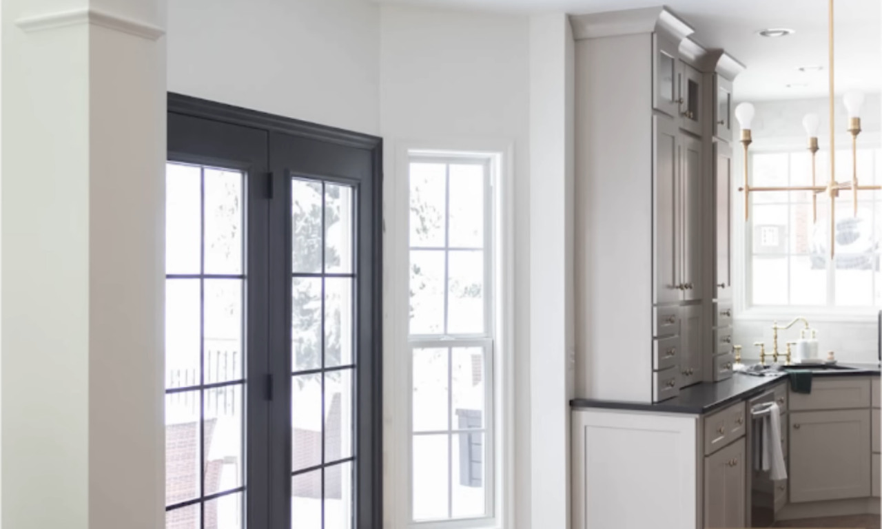 Bright kitchen with white walls, tall gray cabinets, black countertops, and large black-framed glass doors letting in natural light, with a window above the sink.