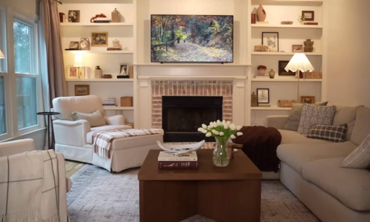 A cozy living room with a neutral color palette featuring a large beige sectional sofa, an armchair with a plaid throw, and a wooden coffee table topped with white tulips and books. Built-in shelves surround a brick fireplace with a flat-screen TV above it displaying a nature painting. The shelves are decorated with books, framed photos, and pottery, adding warmth and personality to the space.