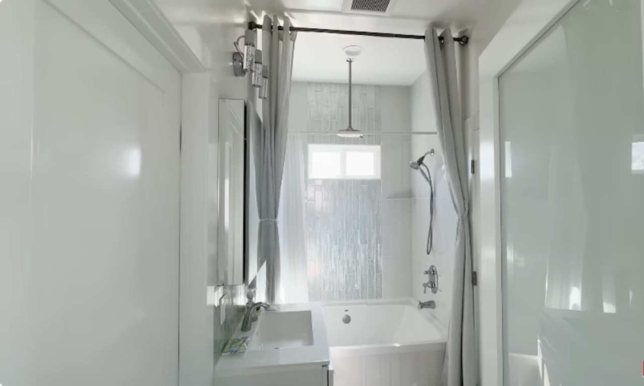 A sleek and modern bathroom with a white vanity and a bathtub-shower combo. Gray curtains frame the tub, and a rain shower head is mounted on the ceiling. The walls are covered in glossy white and light gray vertical tiles, and a frosted glass door adds privacy. Natural light filters in from a small window above the tub.