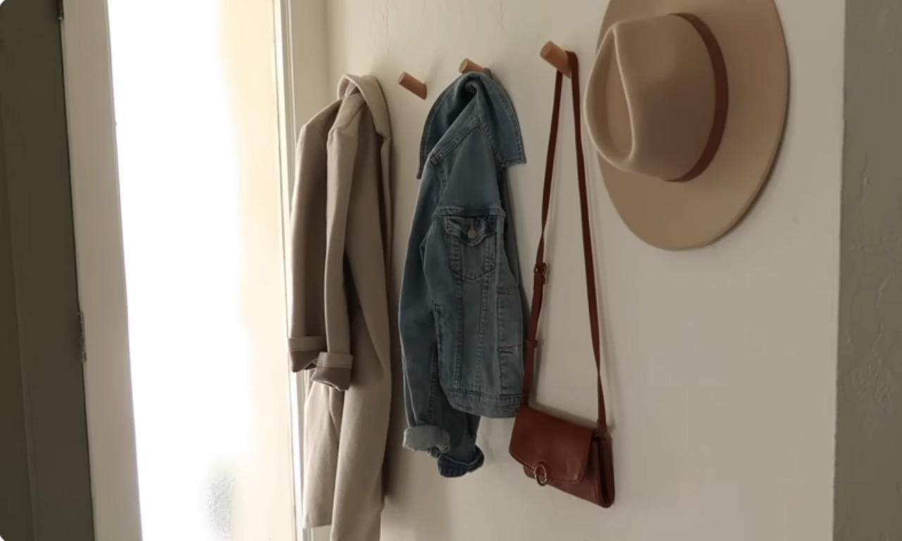 A close-up shot of three minimalist wooden peg hooks mounted on a white wall. A beige trench coat, a denim jacket, and a wide-brimmed hat with a brown leather bag are hanging from the hooks. This shows how to use wall space for storage.