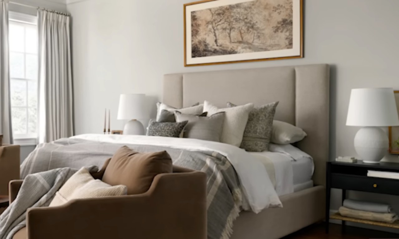A luxurious bedroom featuring a king-sized bed with a tall, neutral-colored upholstered headboard. The bed is dressed in layers of white, light gray, and taupe bedding with a mix of patterned and solid accent pillows. Above the bed hangs a large, framed landscape painting in muted earth tones. In the foreground, the arm of a cozy, brown velvet armchair is visible, suggesting a seating area. A black nightstand with a white lamp is on the right.