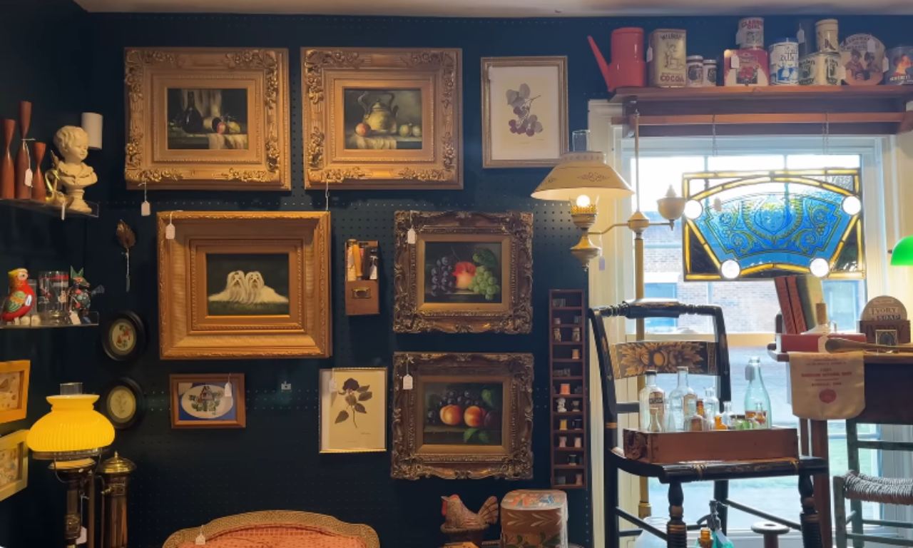 A maximalist, dark gallery wall features a dense collection of oil paintings in ornate gold frames, interspersed with smaller framed art and antique decorative items, creating a cozy and curated vintage aesthetic.