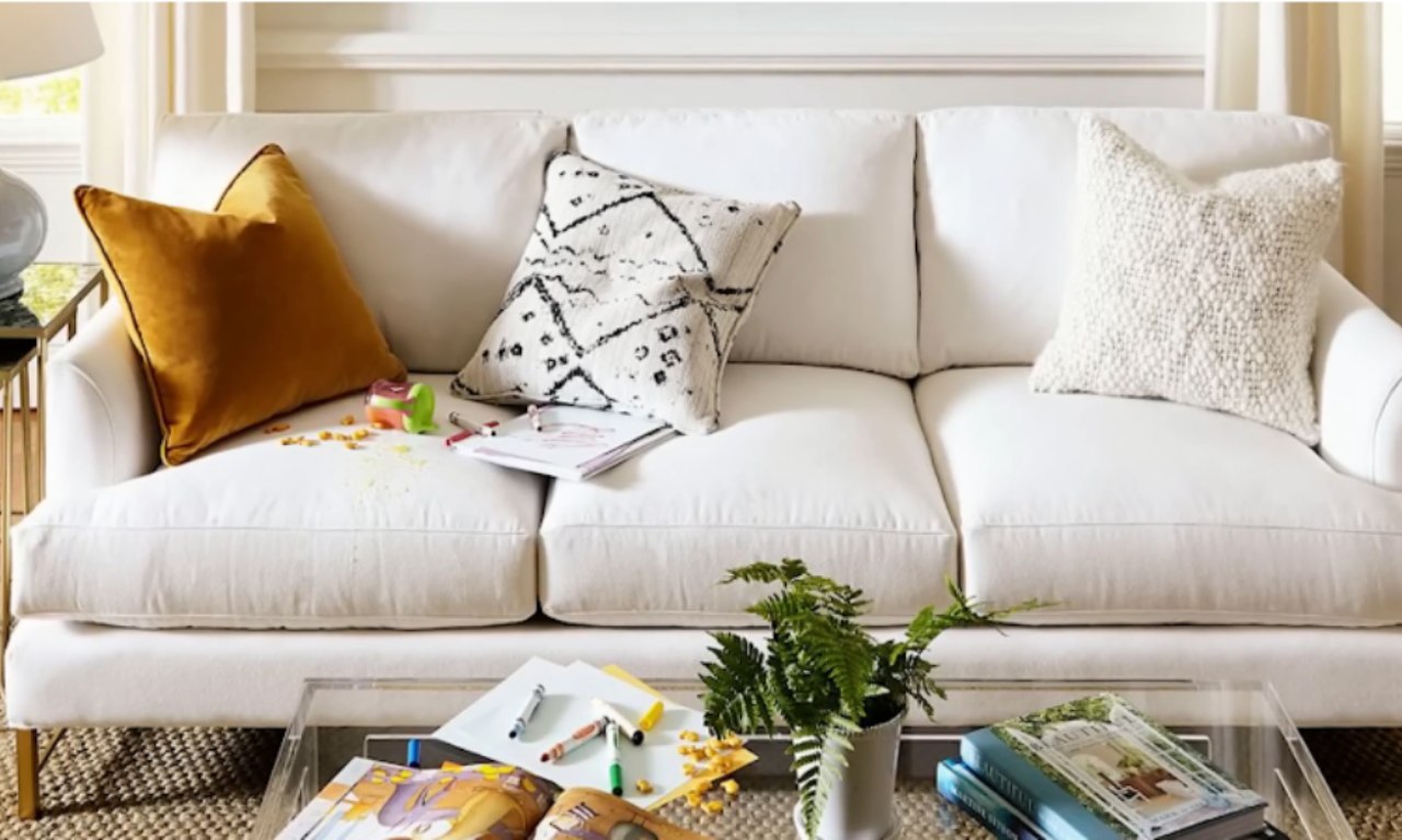 White sofa with three decorative pillows—gold, black-and-white patterned, and cream textured—showing signs of daily use with scattered crayons, papers, and snacks. A glass coffee table in front holds books and a small green potted plant.