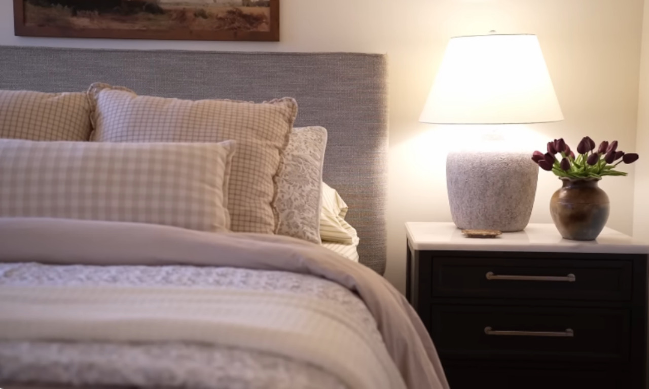 A soft and inviting bedroom scene with a close-up of a bed adorned with layered neutral-toned pillows and bedding in checks and florals. A dark nightstand with brass handles sits beside the bed, holding a textured stone lamp and a rustic vase filled with dark purple tulips. Warm lighting creates a calm and serene ambiance.