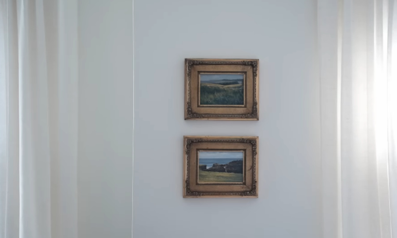 Two small, vintage-style landscape paintings in ornate, gold-toned frames hang vertically on a light-colored wall between two white sheer curtains. The artwork depicts natural scenes with fields and water under cloudy skies.