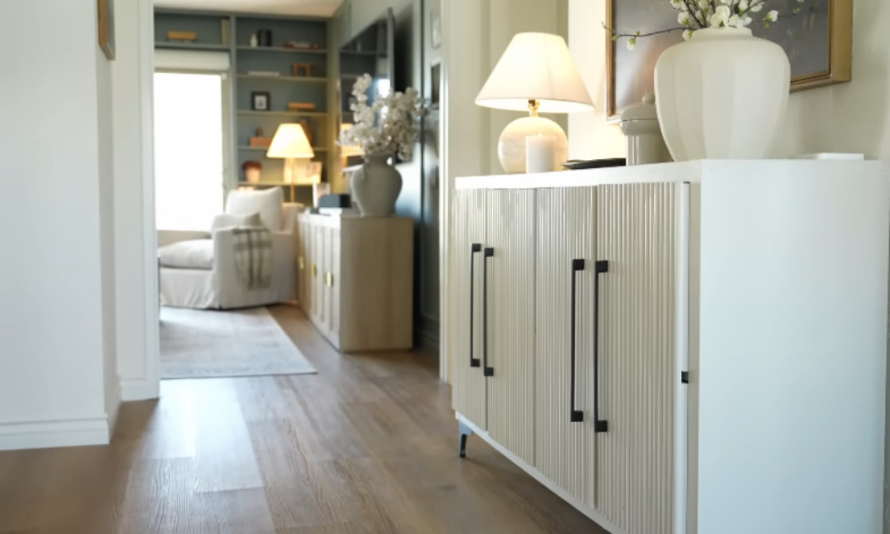 A modern, white console cabinet with vertical ribbing and black rectangular handles. On top of the cabinet are two table lamps, a white cylindrical candle, a white vase with white flowers, and a framed painting. The view extends past the cabinet into a living room with green built-in shelves, a cream-colored armchair, and a second console cabinet. The flooring is light wood laminate.