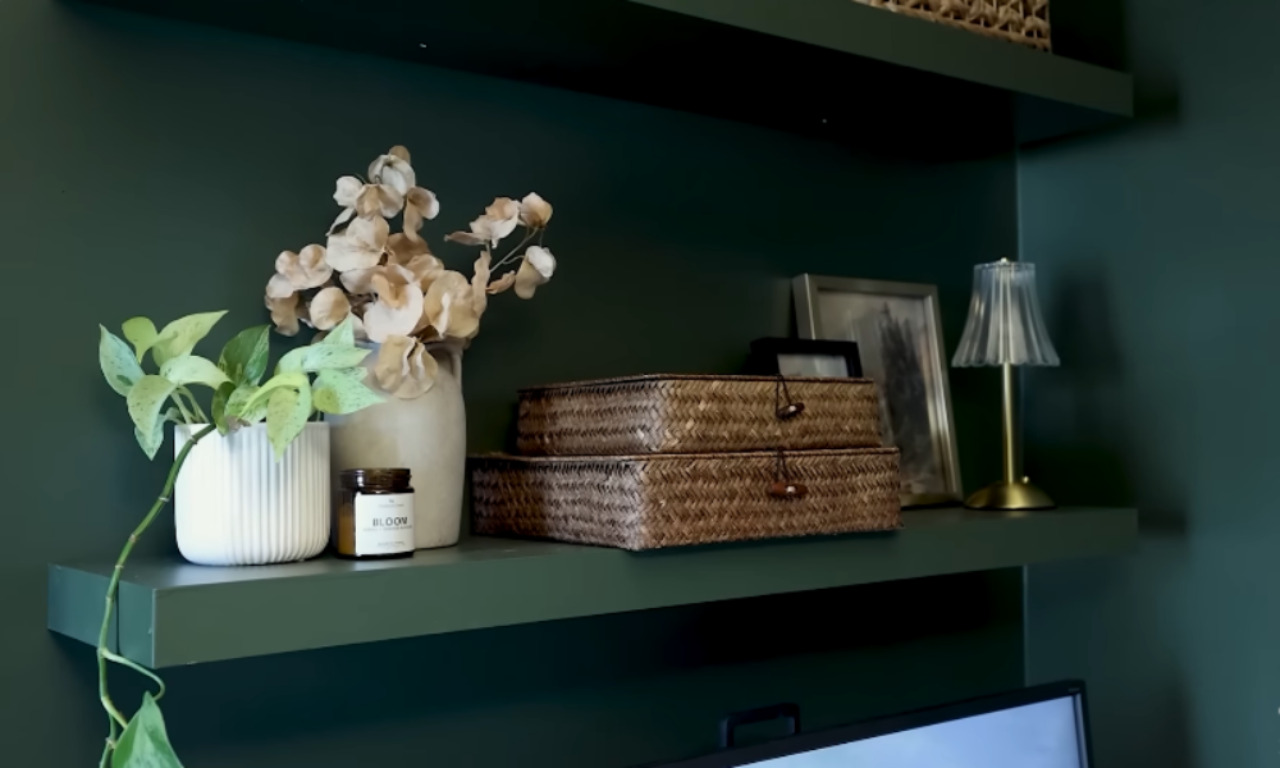 A close-up of a dark green wall-mounted shelf featuring a variety of decorative items, including a trailing pothos plant in a white ribbed pot, dried beige flowers in a ceramic vase, a small amber candle labeled "BLOOM," two stacked woven baskets, framed photos, and a small gold table lamp with a pleated shade.