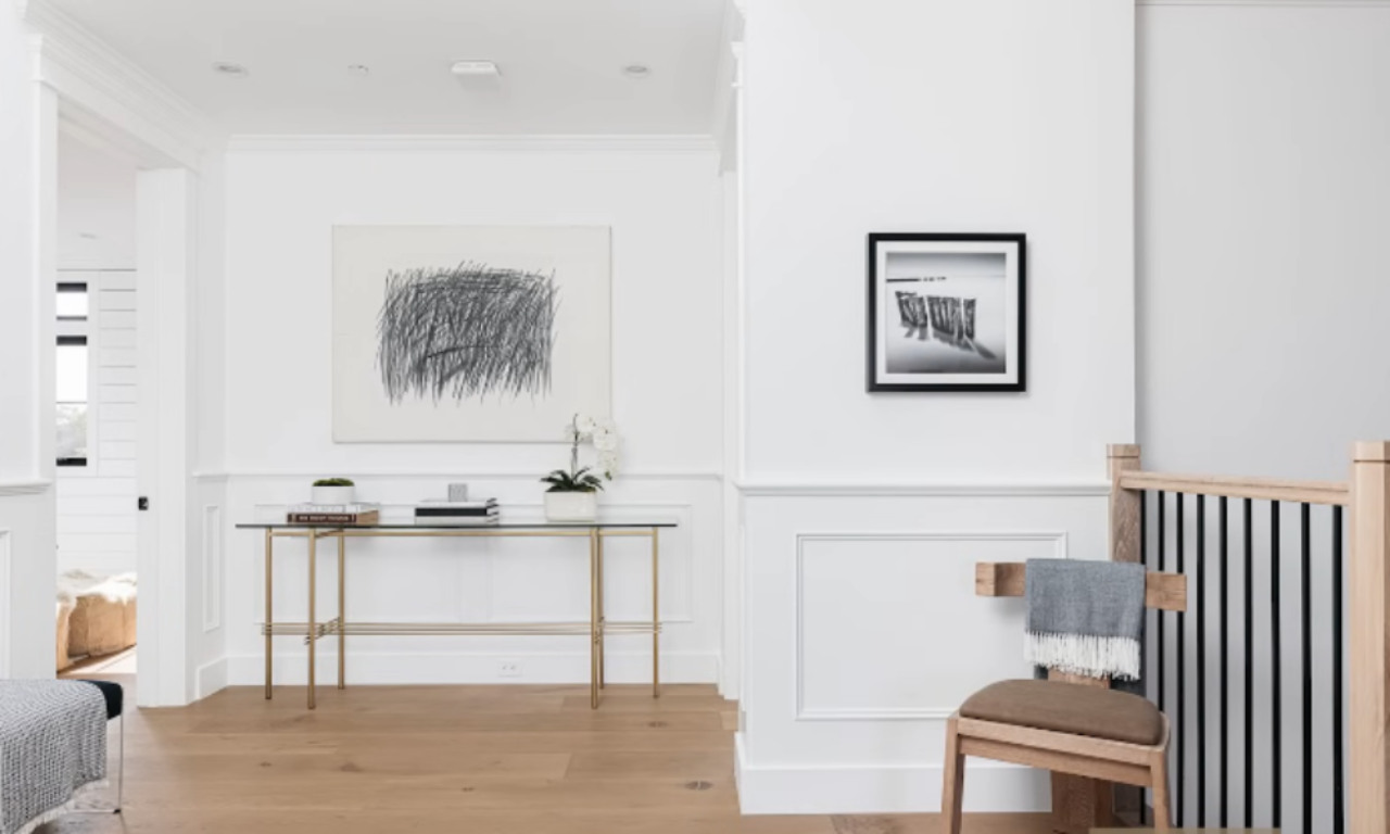 Minimalist hallway with light wood flooring, a modern glass console table against the wall, abstract artwork above it, a framed black-and-white photo on the side wall, and a wooden chair draped with a gray throw.