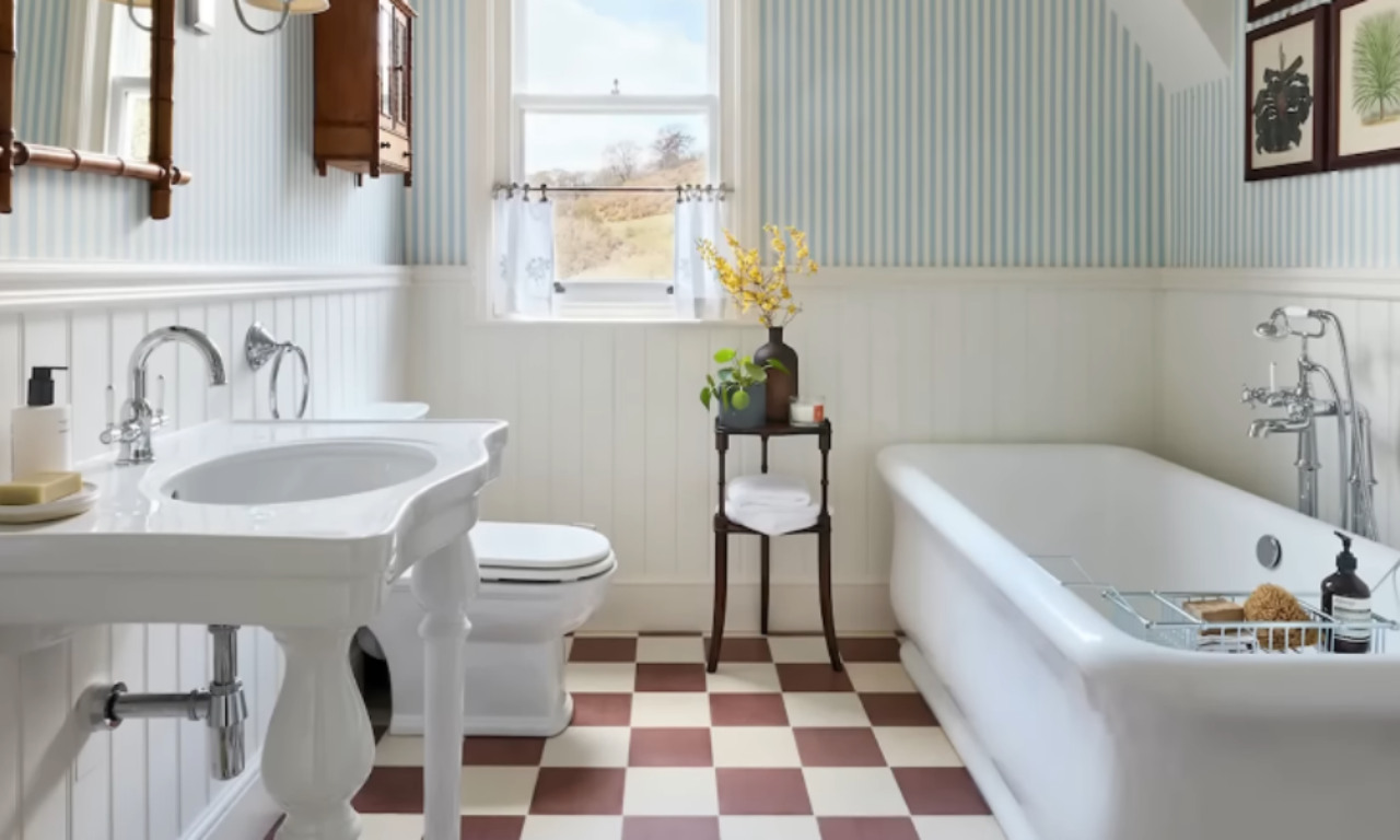 A vintage-style bathroom with a classic white pedestal sink, a white toilet, and a freestanding bathtub. The floor features red and cream checkered tiles, while the walls are decorated with vertical blue and white striped wallpaper above white beadboard paneling. A wooden cabinet, decorative mirror, and a small side table with flowers and towels add charm to the room.