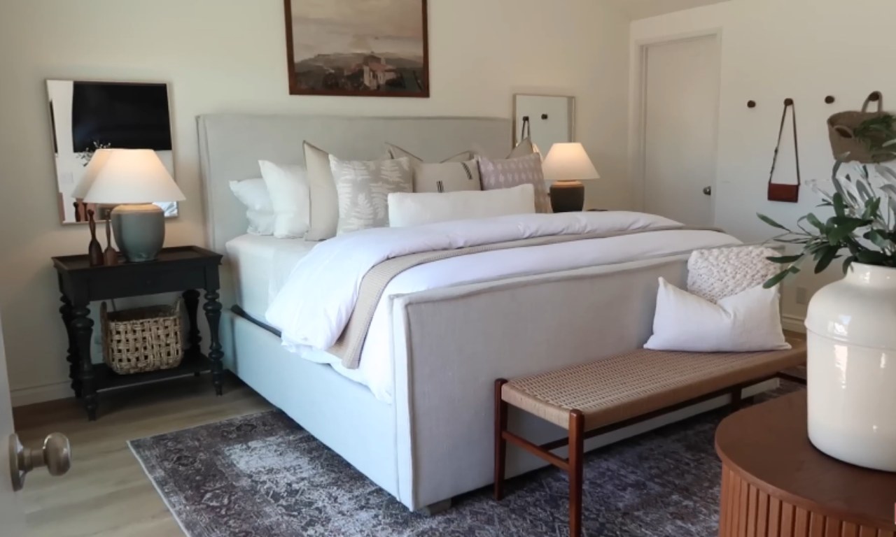 A bright, modern, and cozy bedroom featuring a large, light-grey upholstered bed with white and patterned pillows. A dark, ornate nightstand with a lamp sits on the left, and a wooden bench with a cane seat and a white pillow is placed at the foot of the bed. A patterned area rug is on a light wood floor.